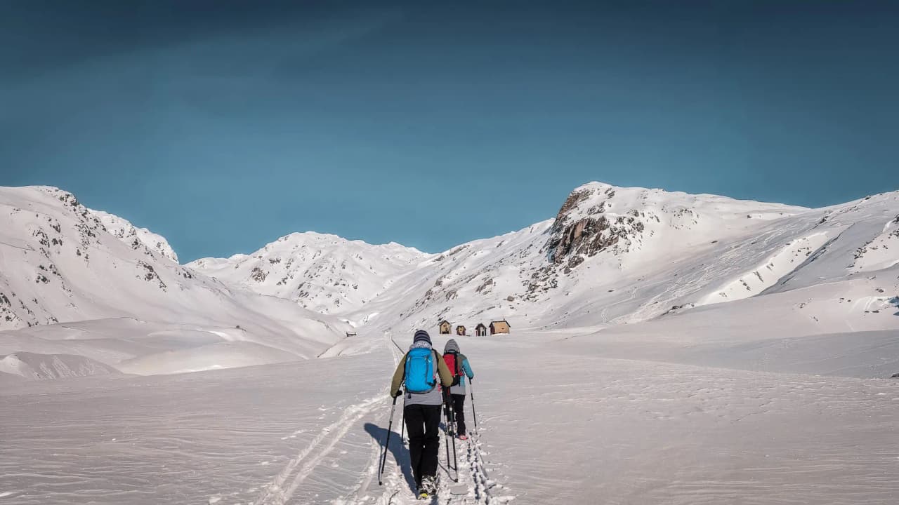Hikers on a snow-covered path, with mountain huts in the distance beneath a brilliant blue sky.