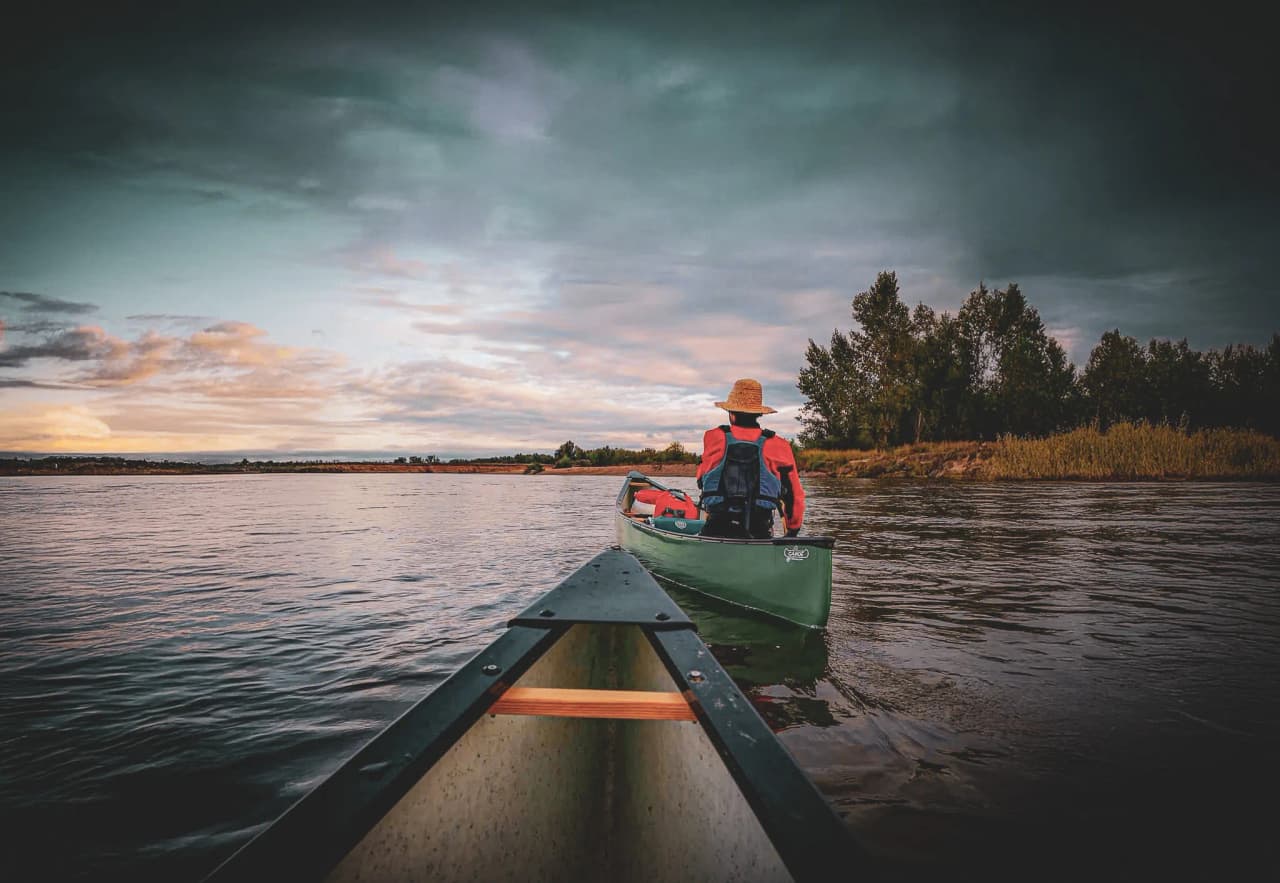 Een vredig waterlandschap, met een persoon in een kano, zijn rug naar de camera, peddel in de hand. Deze persoon draagt een strooien hoed, een rode jas en een reddingsvest. De ar
