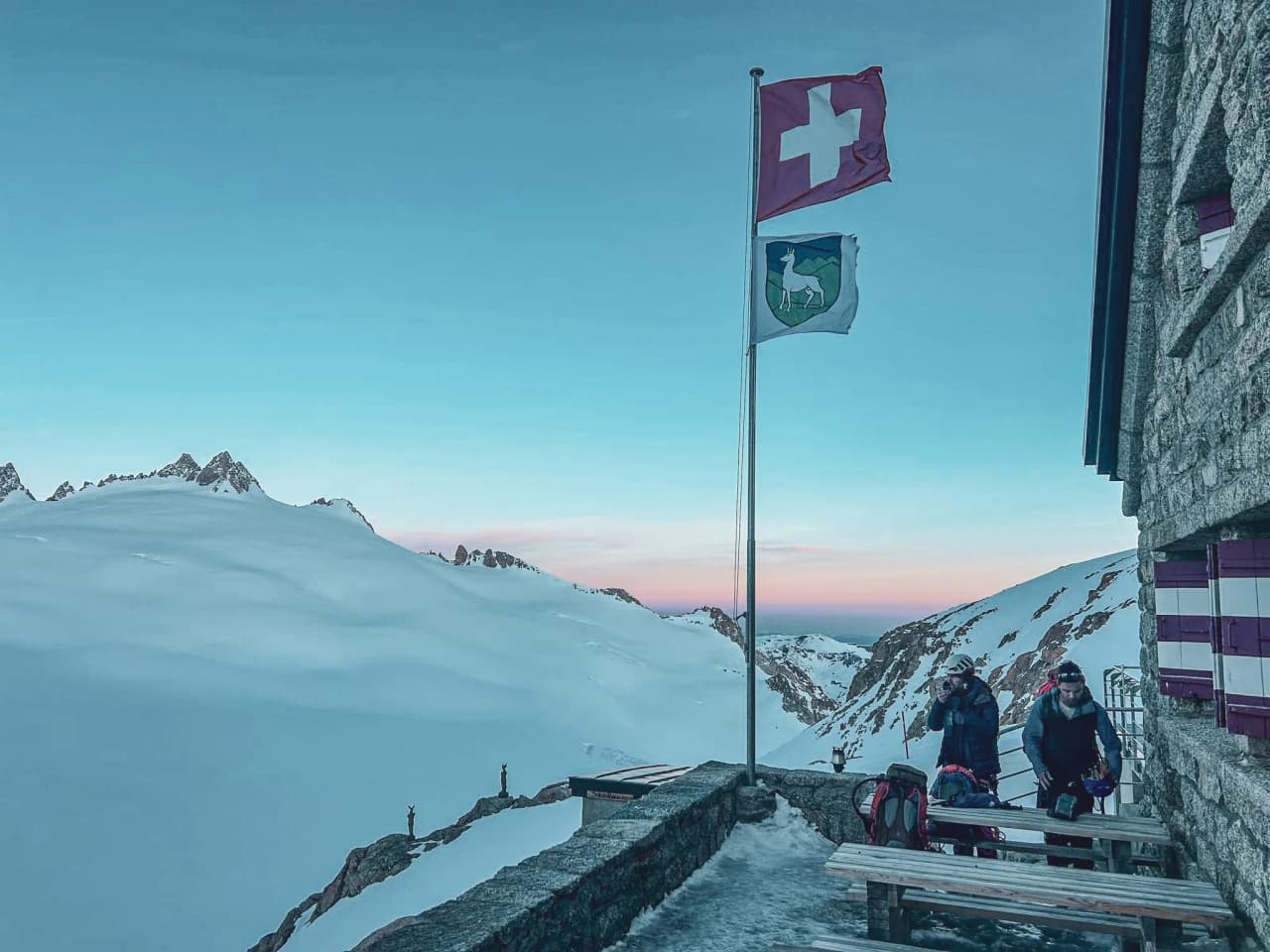 Terrace of an alpine Mountain hut, flags flying, icy landscapes under a pastel sky. A mountain escape.