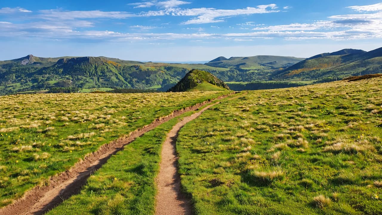 Sentier sinueux à travers des prairies verdoyantes, avec des montagnes en toile de fond sous un ciel lumineux.