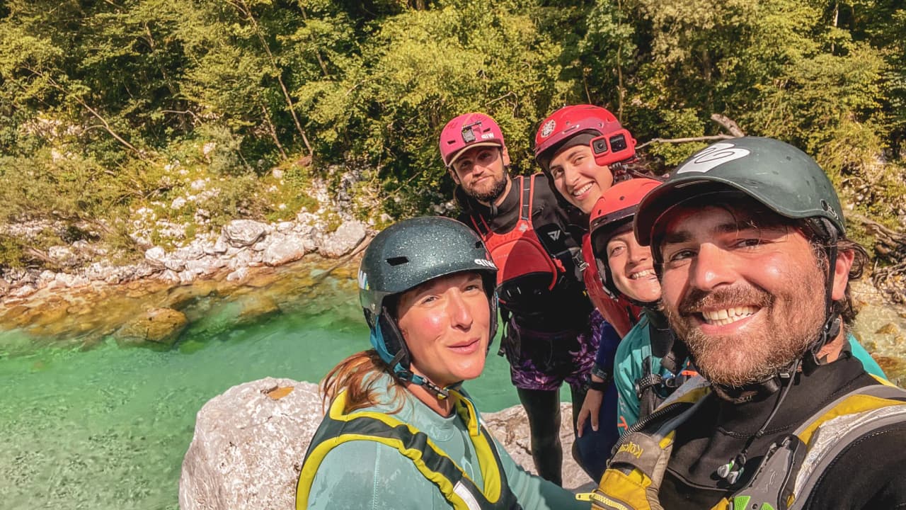Five smiling adventurers pose in a packraft on the banks of a crystal-clear Alpine river.