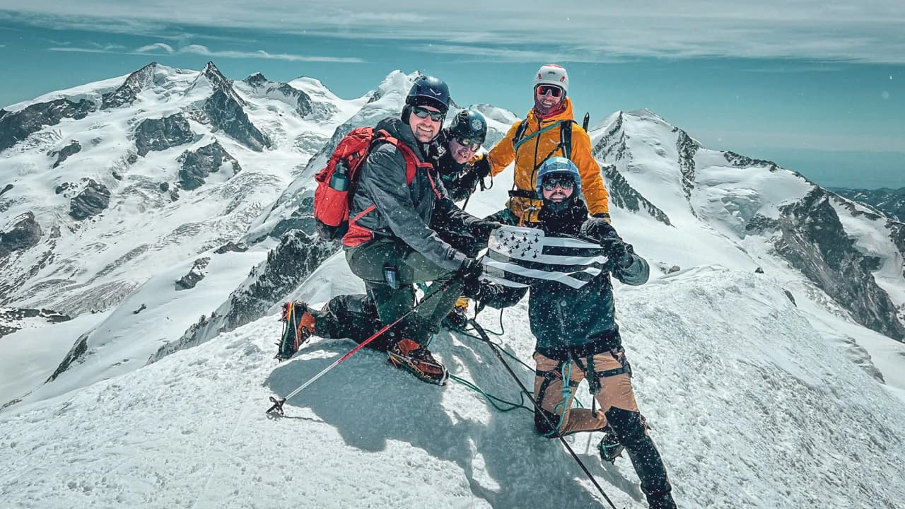 A group of four mountaineers posing on the snow-capped summit of Mont Blanc, with majestic scenery in the background.