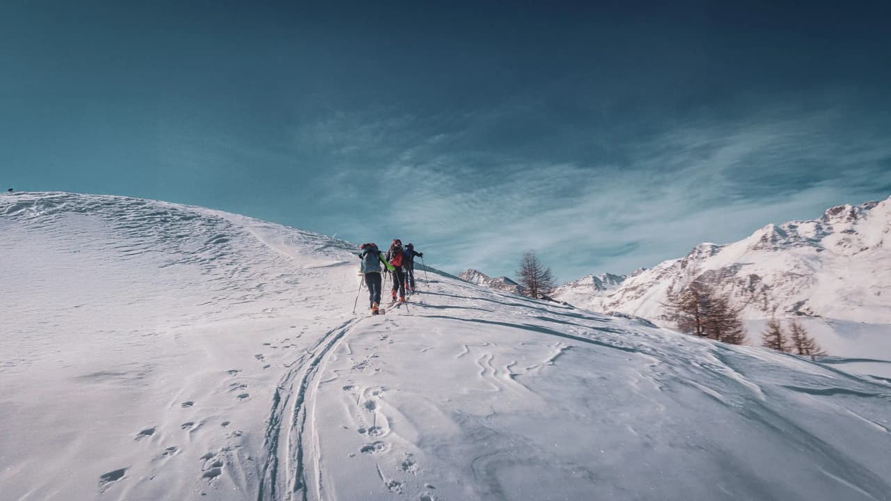 Groupe de randonneurs avançant sur un versant neigeux avec des sommets alpins en arrière-plan.