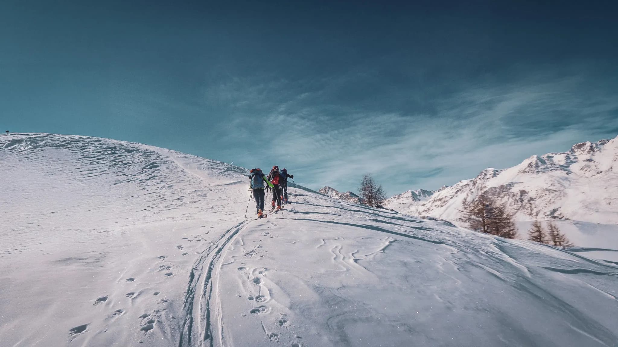 Groupe de randonneurs avançant sur un versant neigeux avec des sommets alpins en arrière-plan.