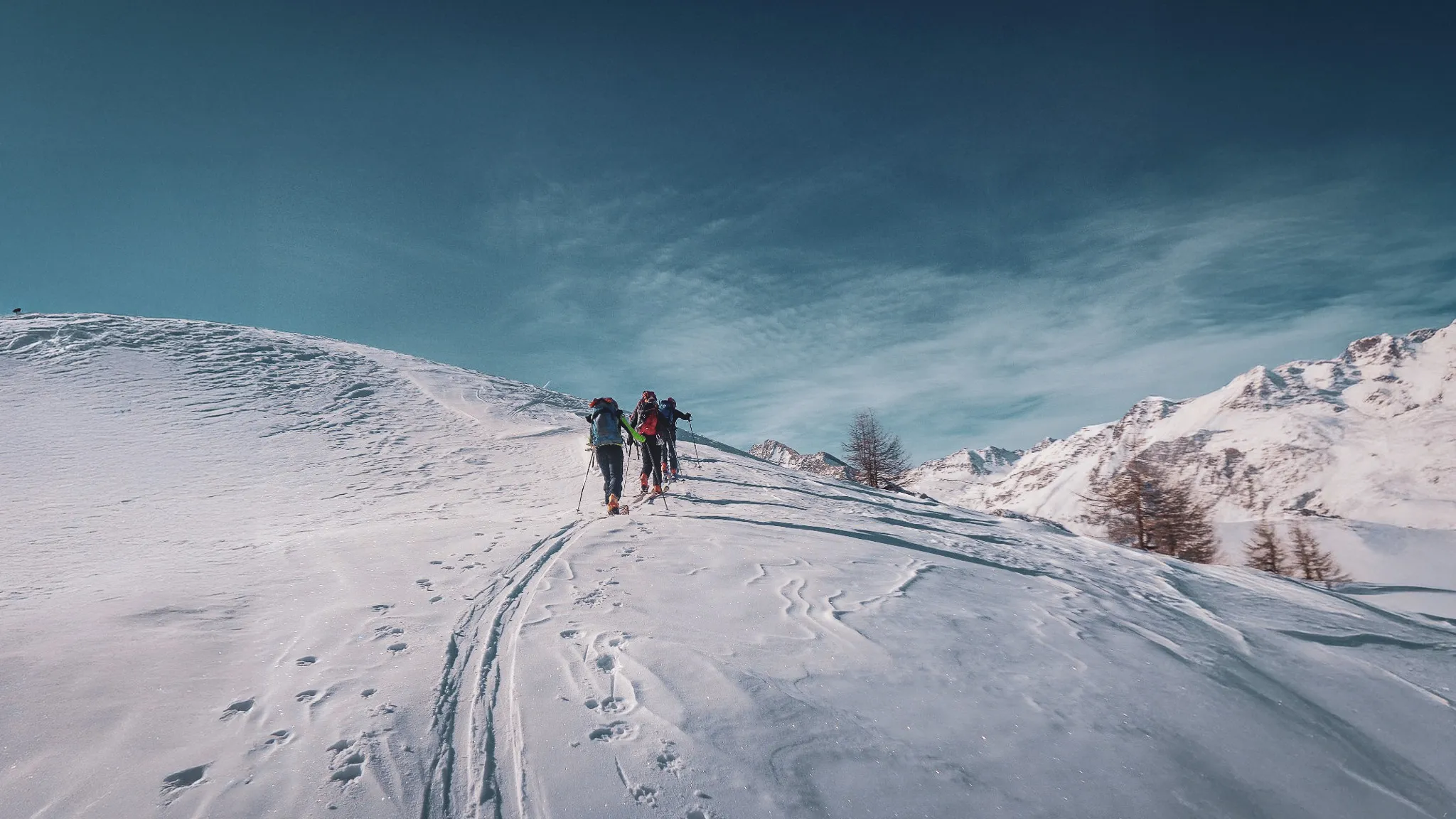 Groupe de randonneurs avançant sur un versant neigeux avec des sommets alpins en arrière-plan.