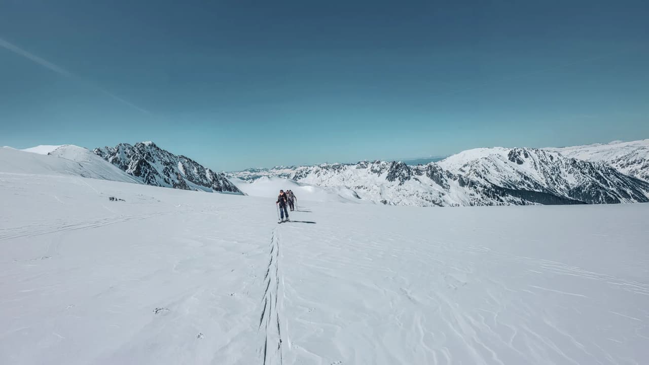 Three skiers cross a pristine glacier, surrounded by snow-capped mountains under a blue sky.