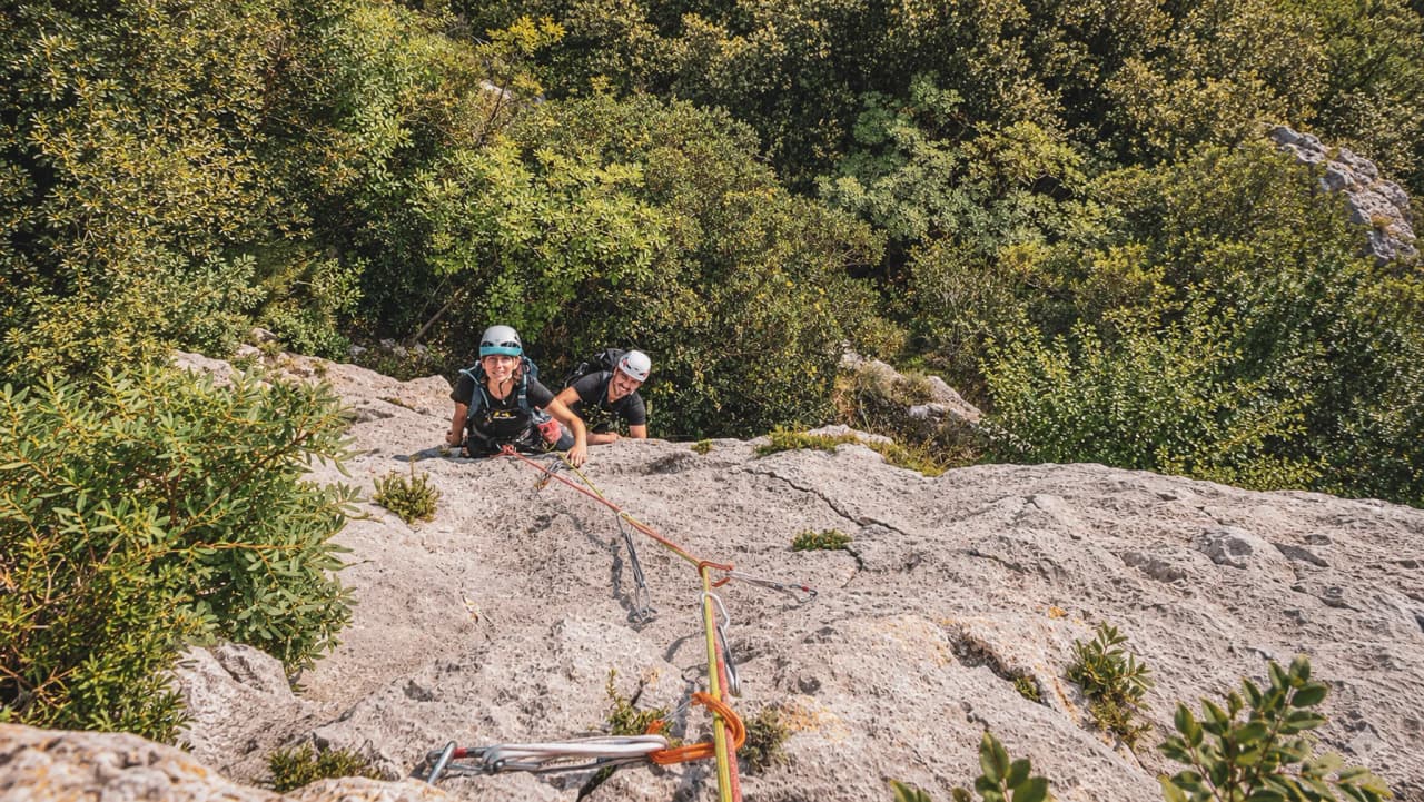 Two climbers are making their way up a rock face, surrounded by greenery, under a sunny sky.