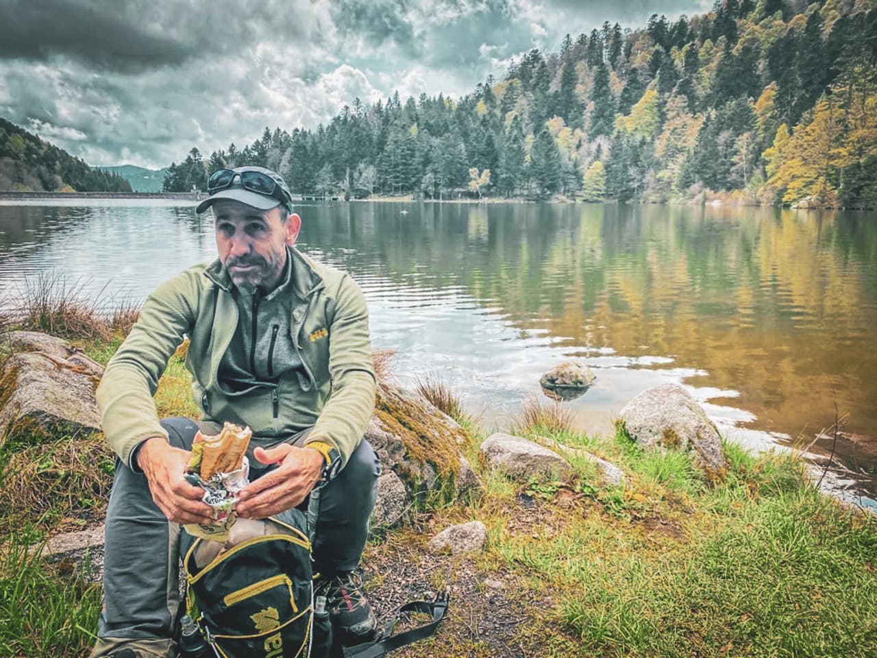 A man sitting by a peaceful lake, surrounded by lush green forests, enjoying a sandwich.