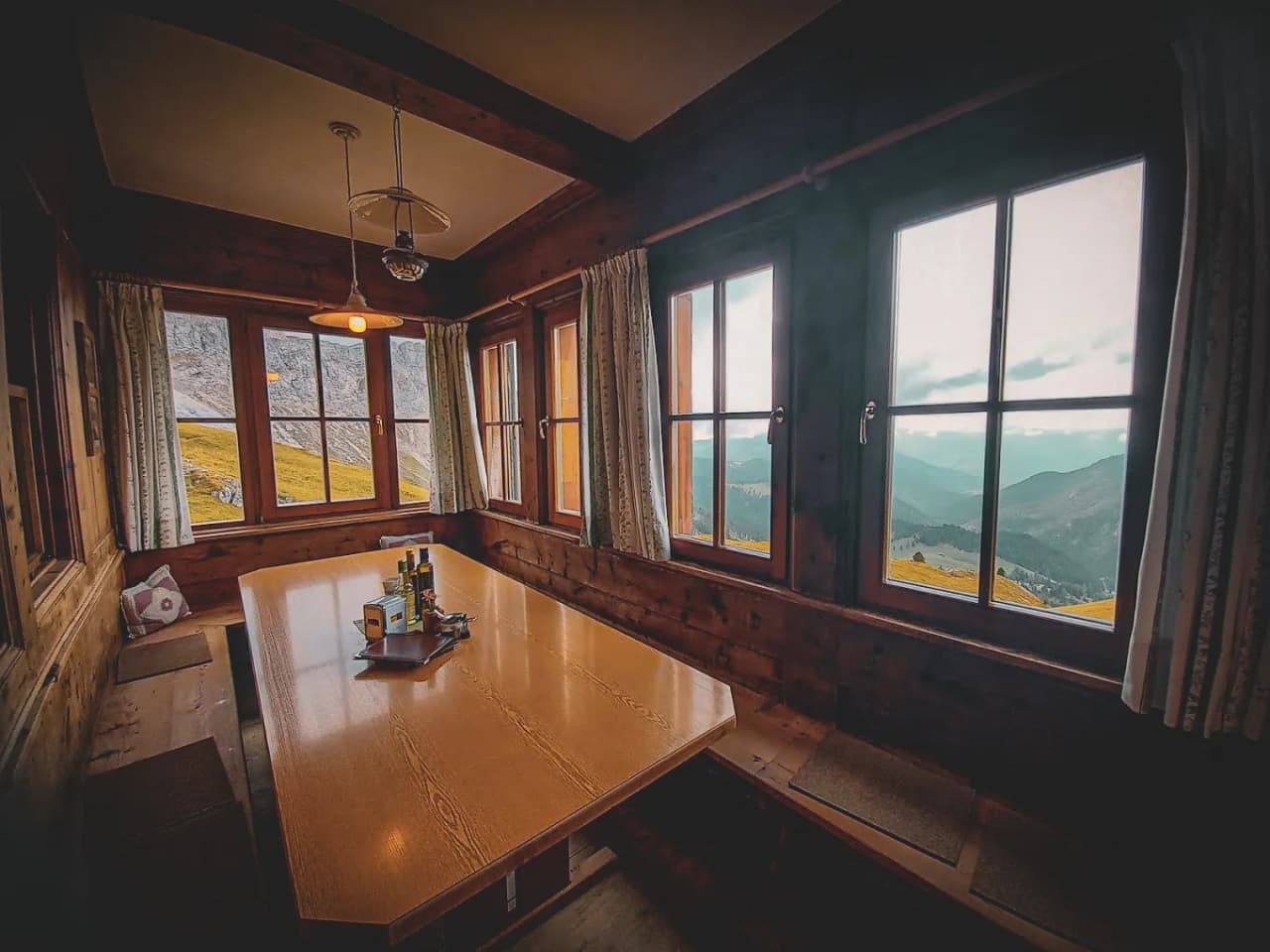 A chalet interior with a large wooden table in the centre, surrounded by benches. Through the windows, you can see an impressive mountain landscape, illuminated by natural light. White curtains frame the windows