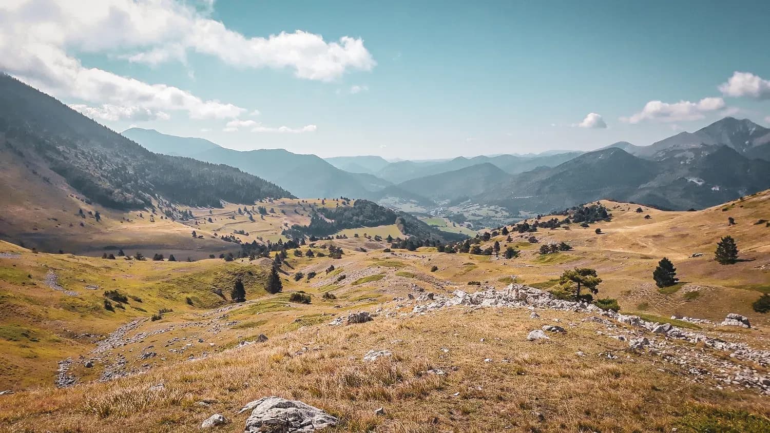 A mountain landscape of golden alpine pastures, blue skies, ridges and lush green valleys.