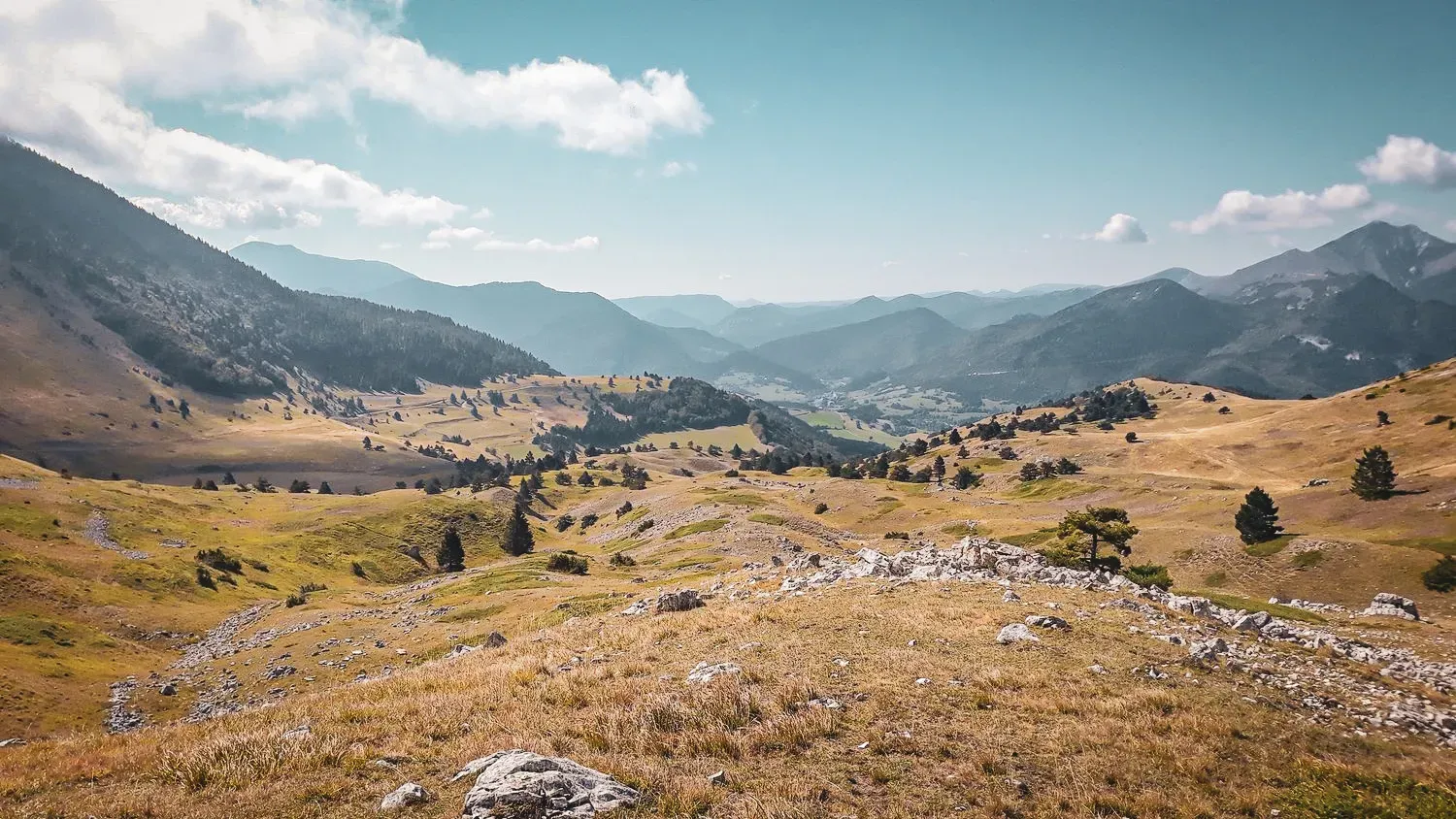 A mountain landscape of golden alpine pastures, blue skies, ridges and lush green valleys.