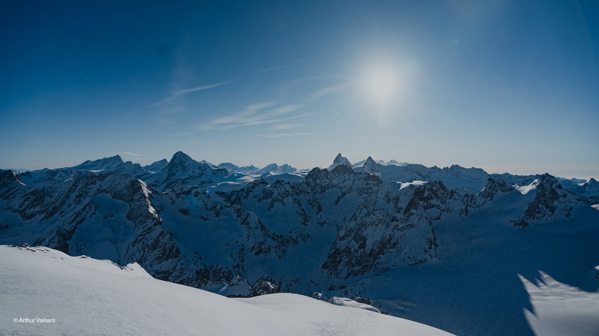 A sweeping alpine panorama under a sunny sky, snow-capped mountains and majestic glaciers.
