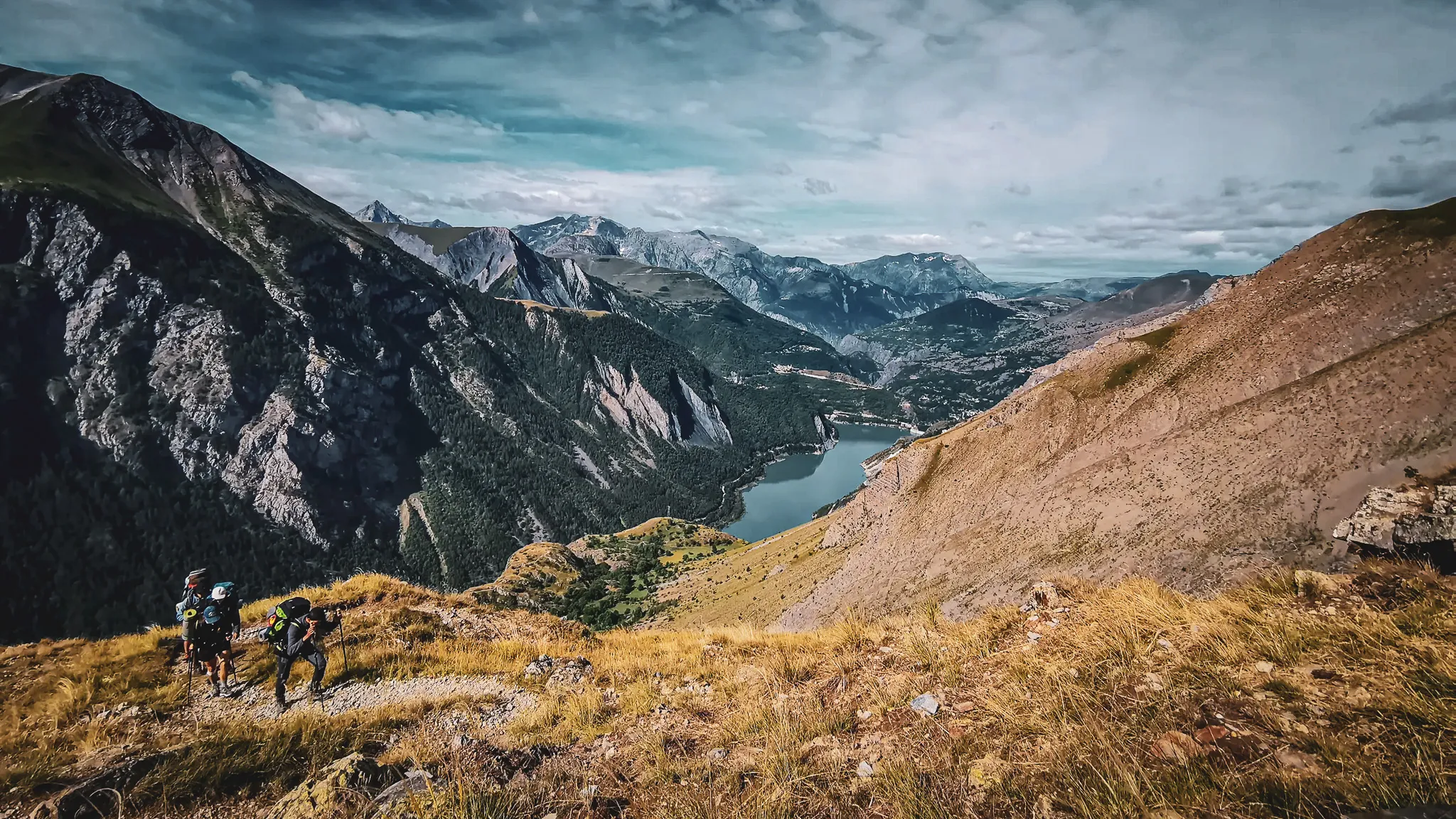 Un aperçu époustouflant des montagnes avec randonneurs, verdure et lac scintillant sous le ciel bleu.