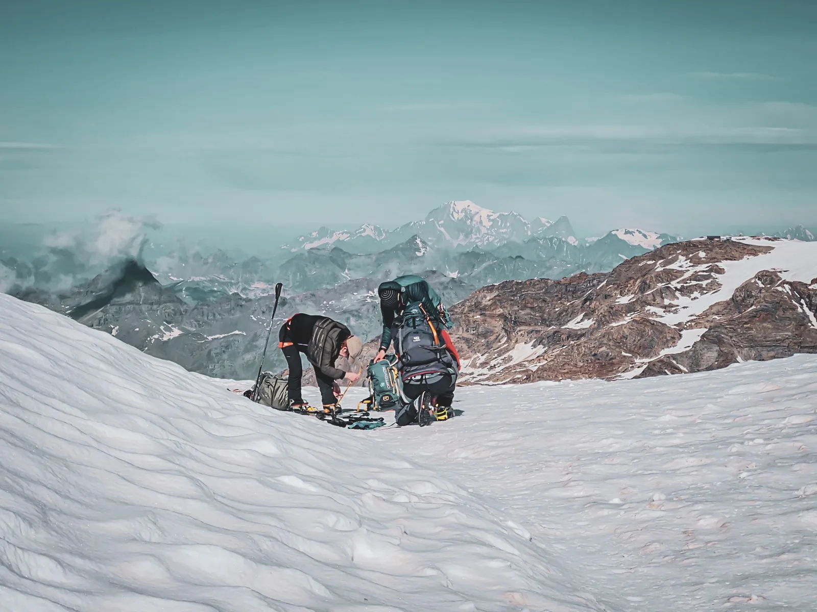 Two mountaineers in full preparation on a glacier, with majestic mountains in the background.