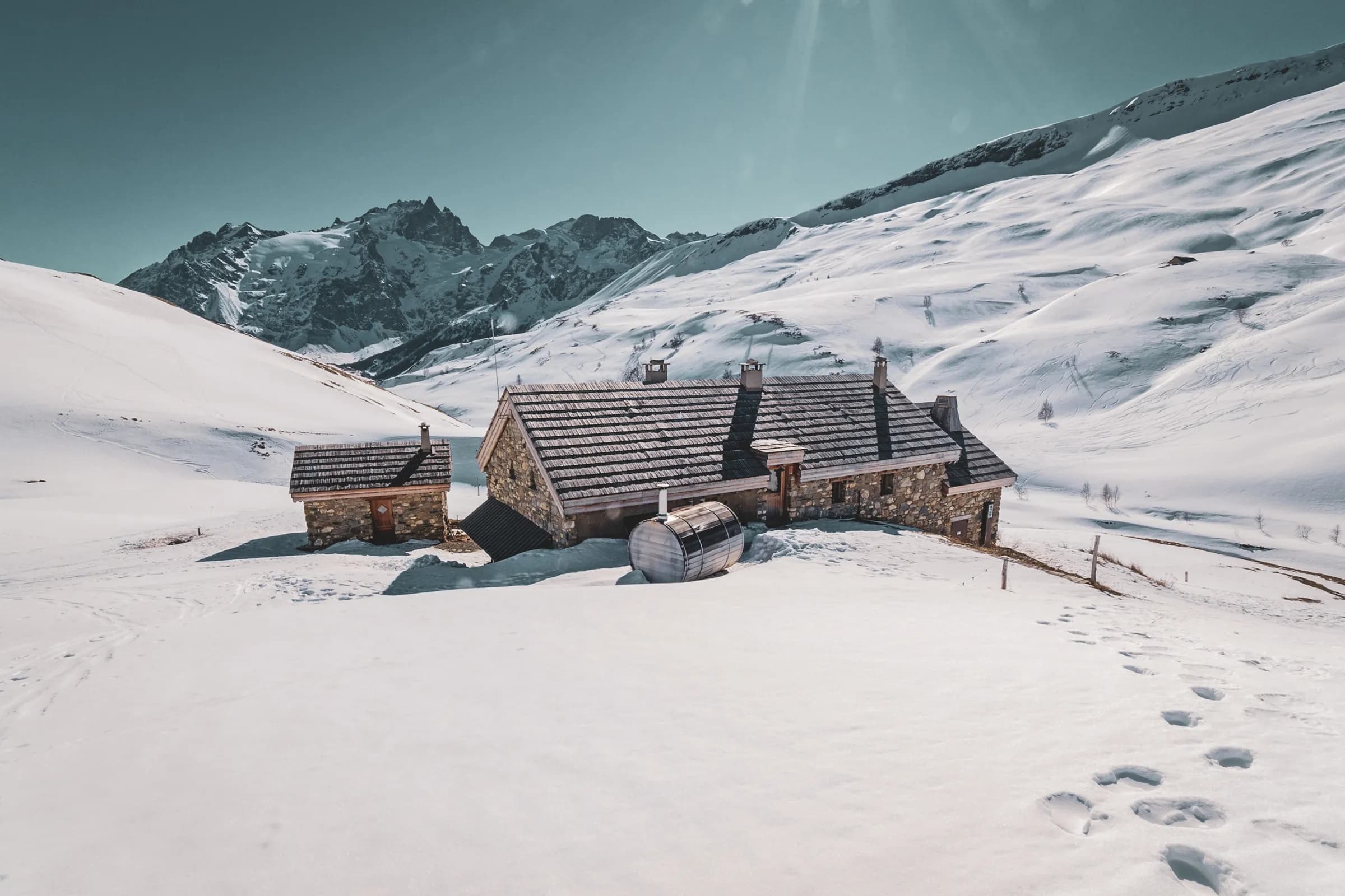 Een stenen chalet, omringd door besneeuwde bergen en strakblauwe luchten.