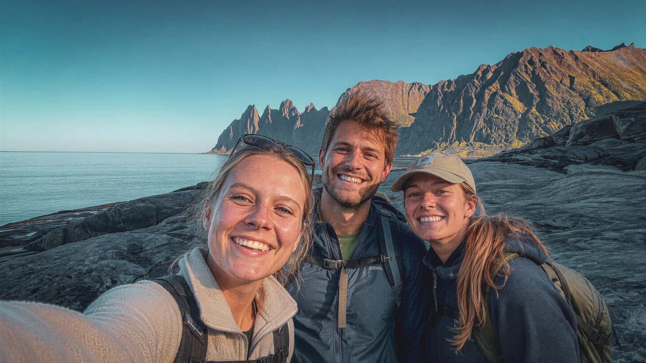 Drie lachende wandelaars aan het water, met majestueuze bergen op de achtergrond.