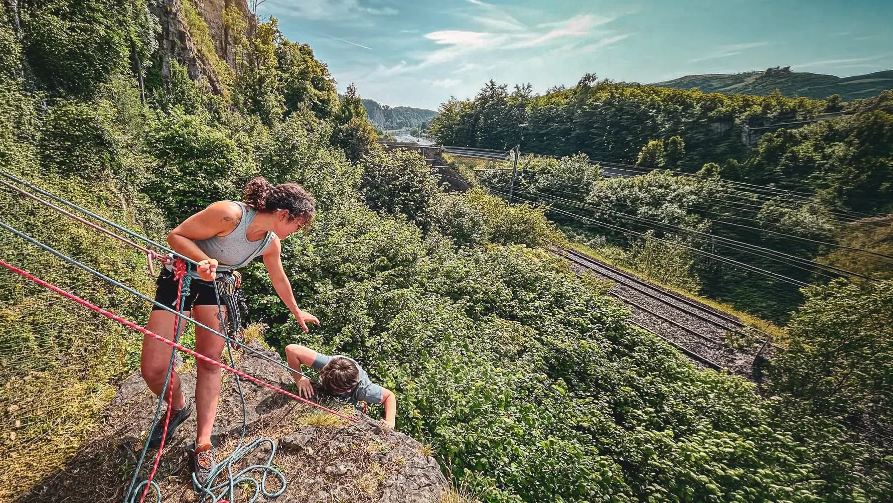 An instructor guides a beginner climber on a green cliff in Yvoir, Belgium.