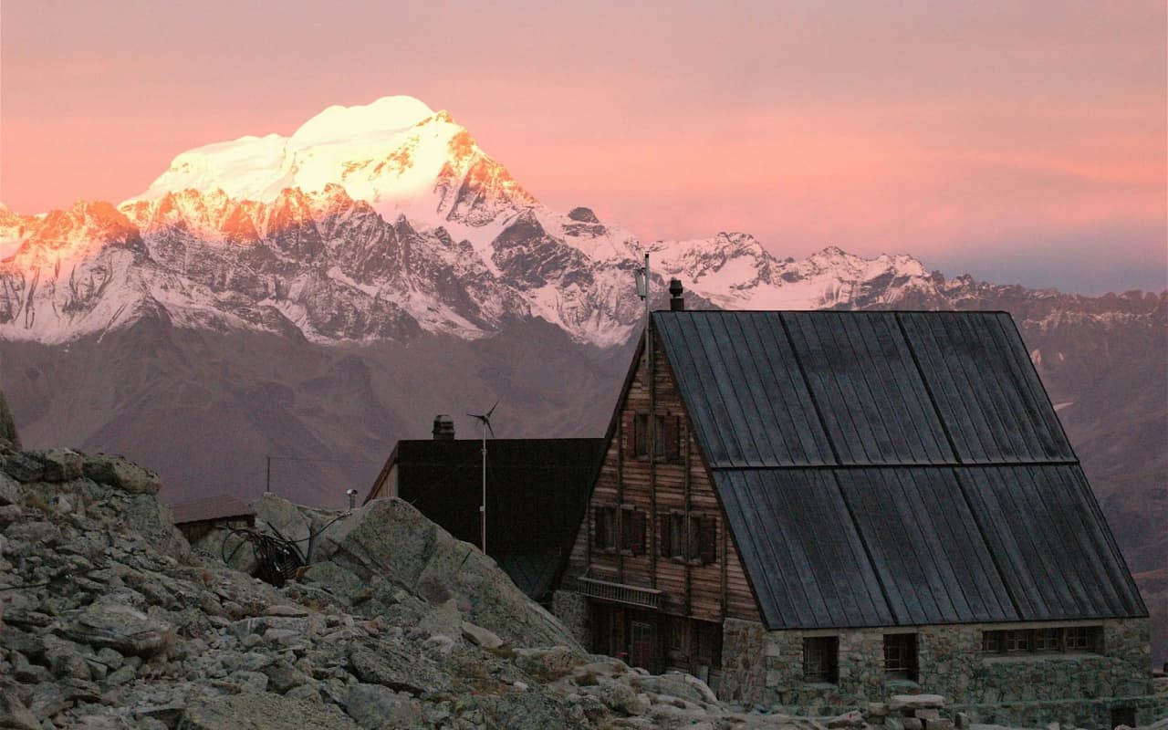 Panoramic views of snow-capped peaks at sunrise, with a picturesque Mountain hut in the foreground.